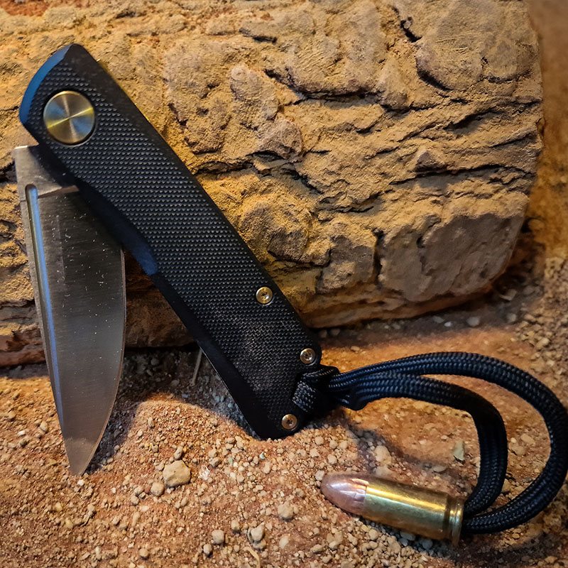 The owners Real Steel Luna pocket knife on a wooden log in a desert like environment. Shows a custom bullet lanyard attached.
